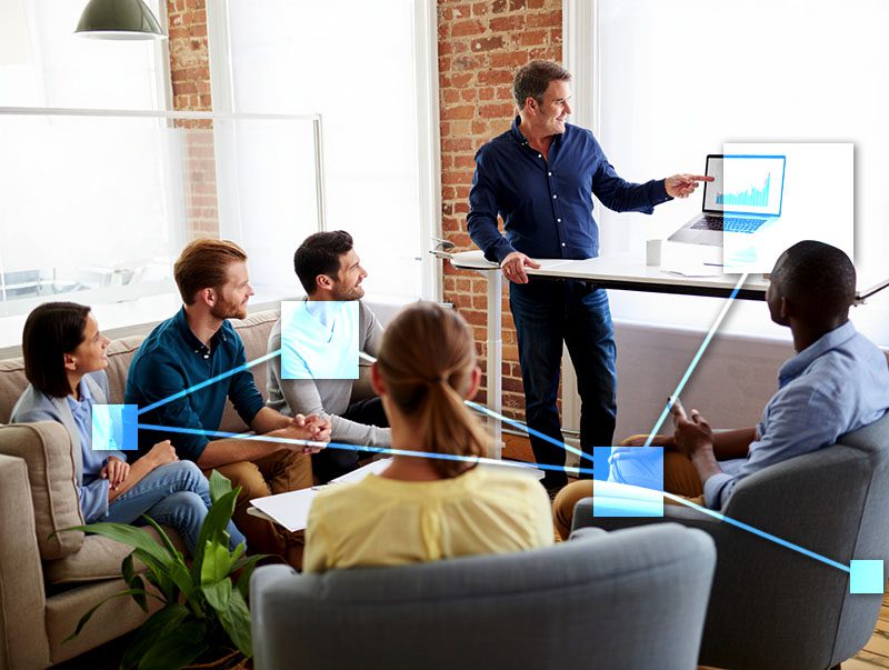 Presenter pointing to laptop in team meeting showing increased sales. 